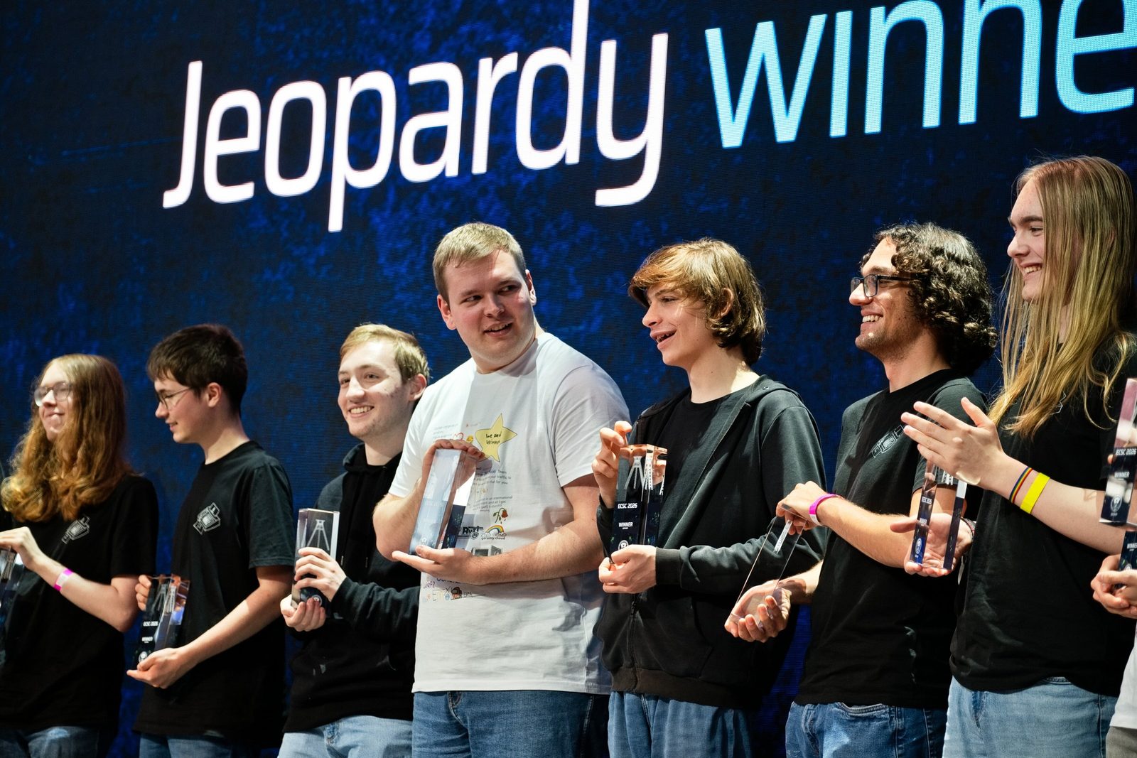 Mitglieder von Team Germany stehen lachend mit gläsernen Trophäen vor der Leinwand „Jeopardy winner“.