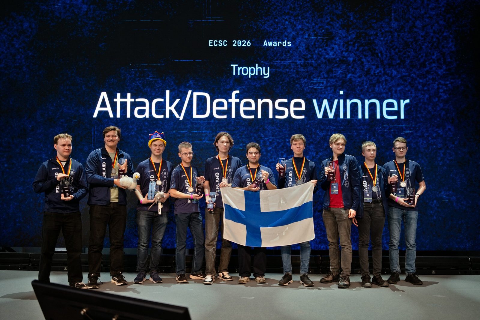 Das finnische Team mit Flagge, Trophäen und Medaillen auf der Bühne vor der Leinwand „Attack/Defense winner“.