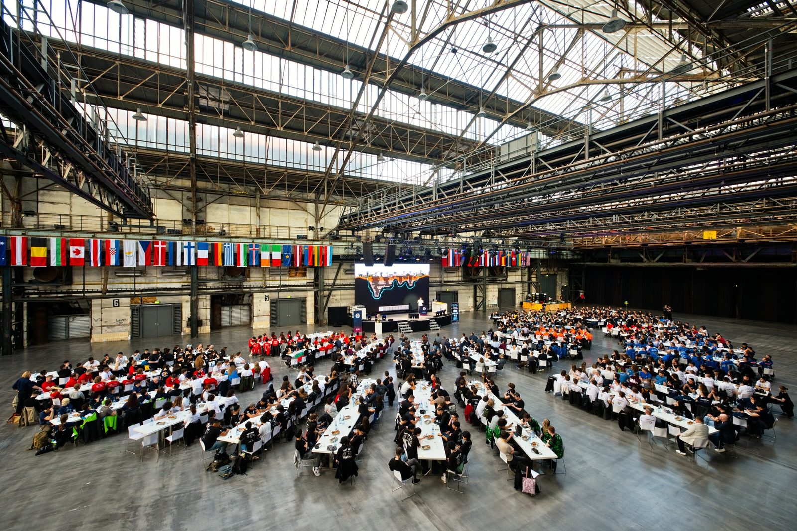 Blick von oben in die Jahrhunderthalle Bochum: Hunderte Teilnehmende an langen Tischen, darüber Flaggen und das Stahl-Glas-Dach.