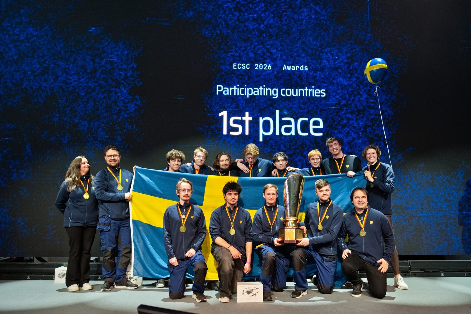 Das schwedische Team mit Flagge, Pokal und Goldmedaillen auf der Bühne vor der Leinwand „1st place“.