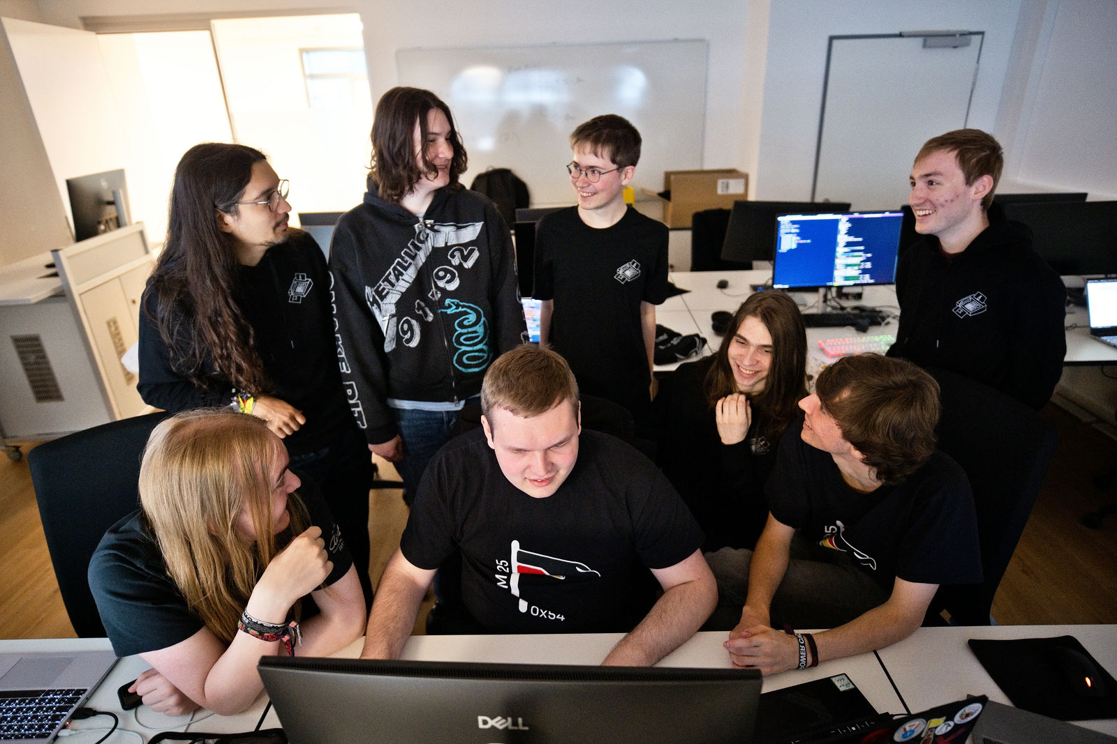 Mehrere Personen sitzen und stehen an einem Arbeitsplatz mit Computern. Sie wenden sich einander zu, sprechen miteinander und lachen.